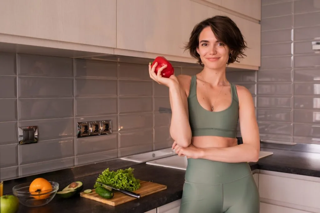 Woman in activewear holding a red pepper in a bright kitchen with fresh vegetables, representing healthy eating for weight management